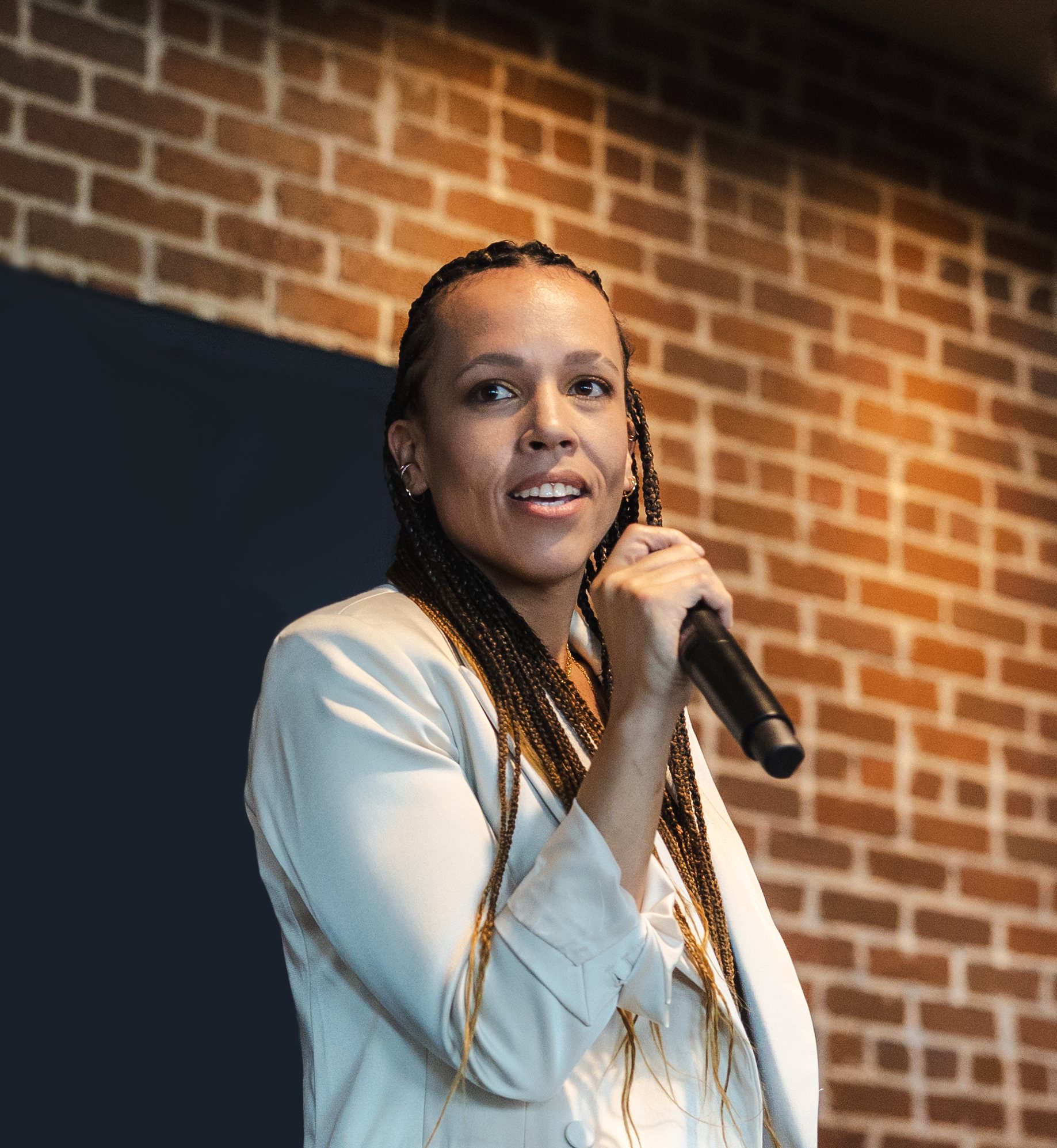 Br33 Jon3s speaking on stage with a microphone, in an ivory blazer in front of a brick wall